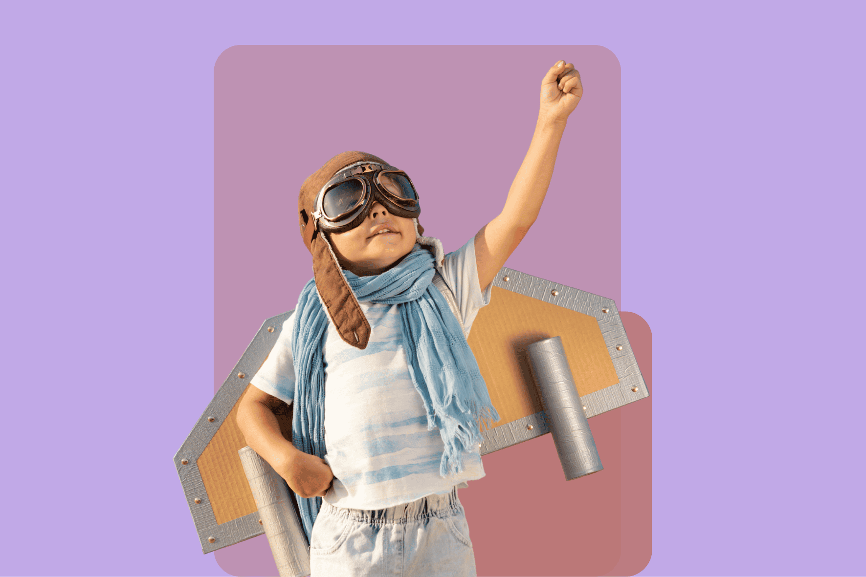 Kleiner Junger mit Fliegerbrille, Fliegermütze und Raketenflügeln aus Pappe, steht vor einem lilafarbenen Hintergrund und streckt seine Faust in die Luft