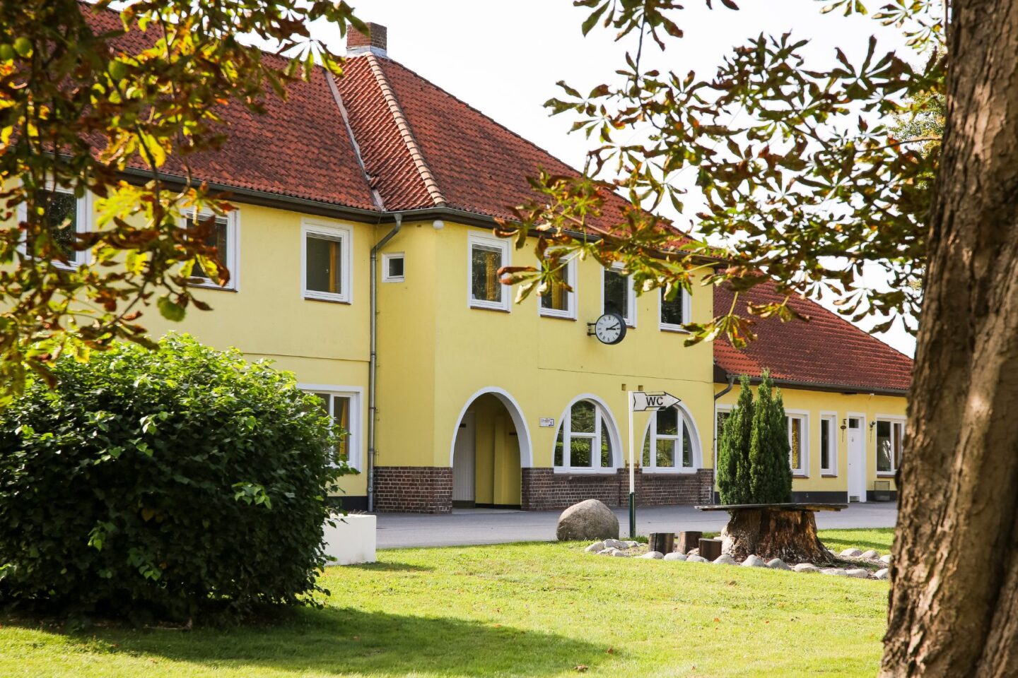 Außenansicht der Freiluftschule Bunthausspitze: Großes gelbes Haus mit rotem Ziegeldach vor einer großen Rasenfläche und Bäumen umringt.