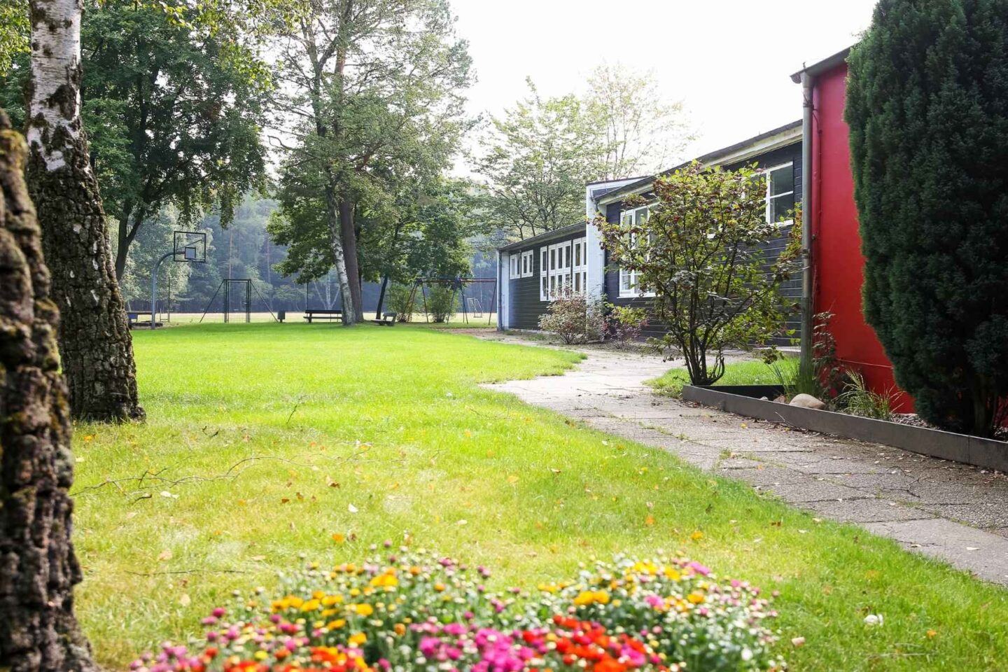 Außenansicht der Freiluftschule Fischbeker Heide: Dunkelgraues Haupthaus vor einer großen, grünen Rasenfläche umringt von vielen Bäumen.