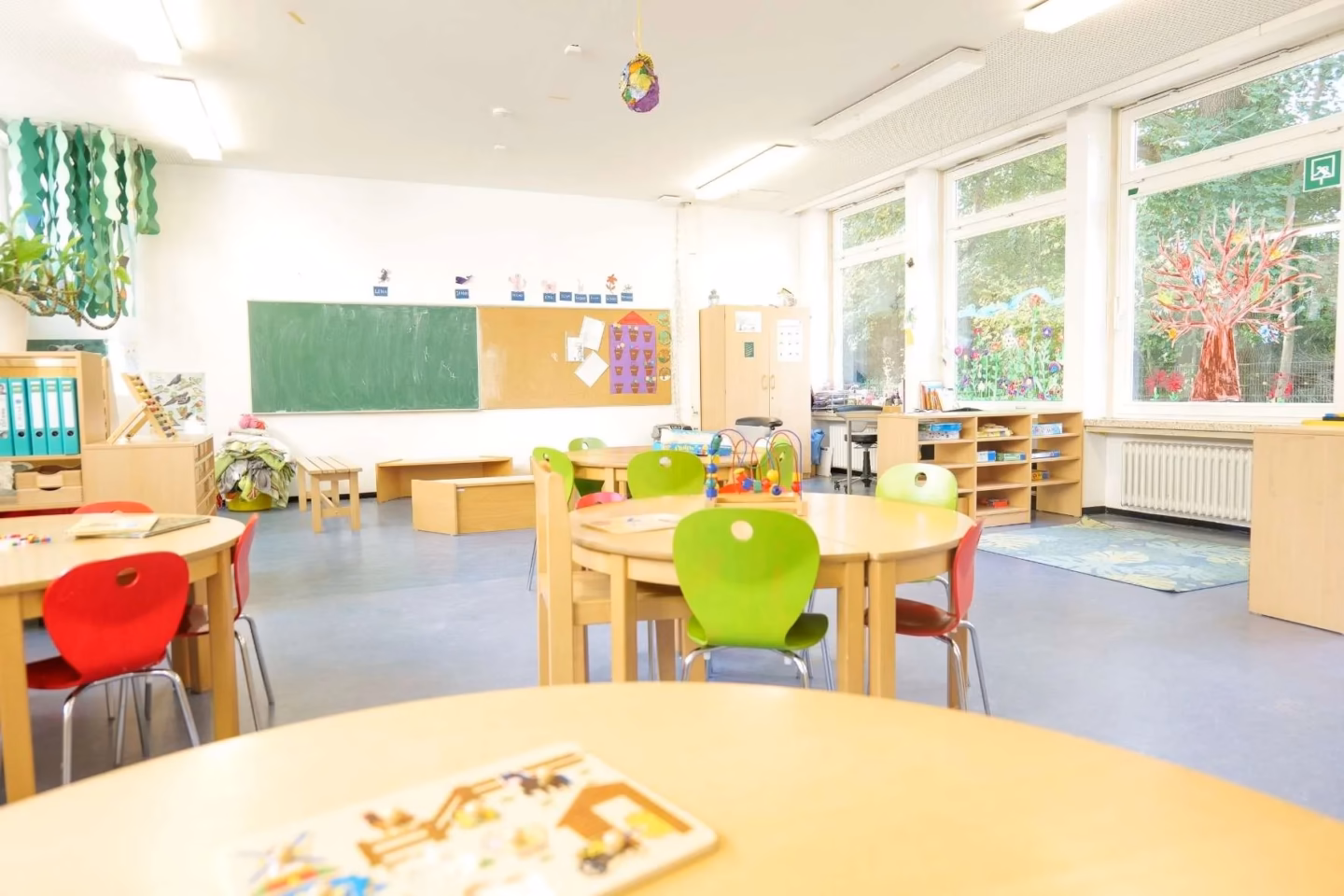 Heller, lichtdurchfluteter Gruppenraum der Kita Redder mit vielen runden Tischgruppen, Spielecken und einer großen grünen Tafel an der Wand.