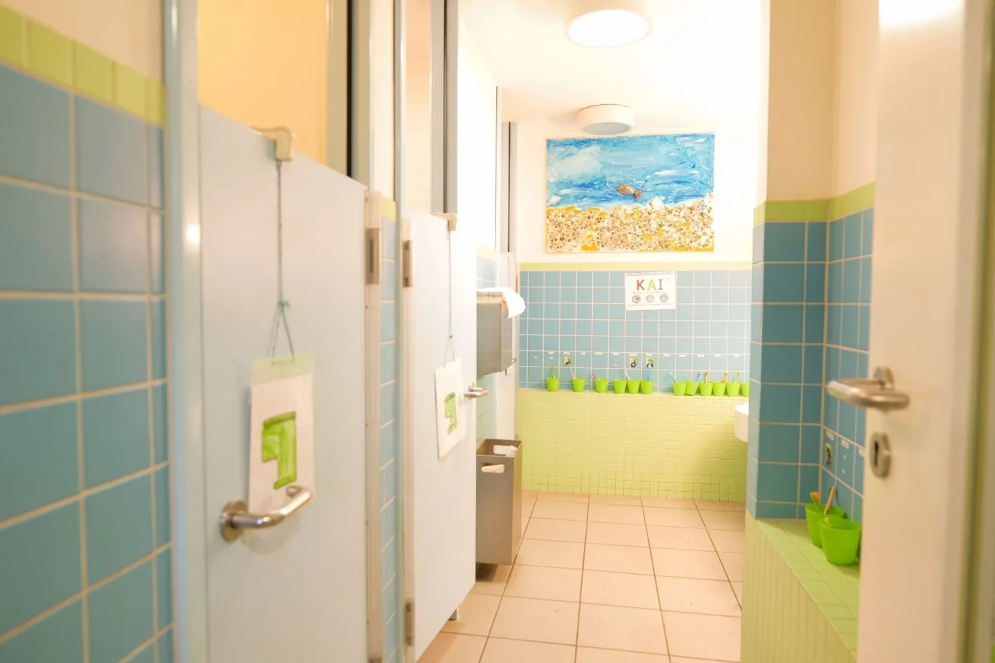 Helles, kindgerechtes Badezimmer in Grüntönen in der Kita Völckersstraße mit Toiletten und, Waschbecken und grünen Zahnputzbechern.